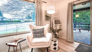 A modern living room with a white armchair, a side table, a lamp, and a view of a balcony with a railing and greenery.