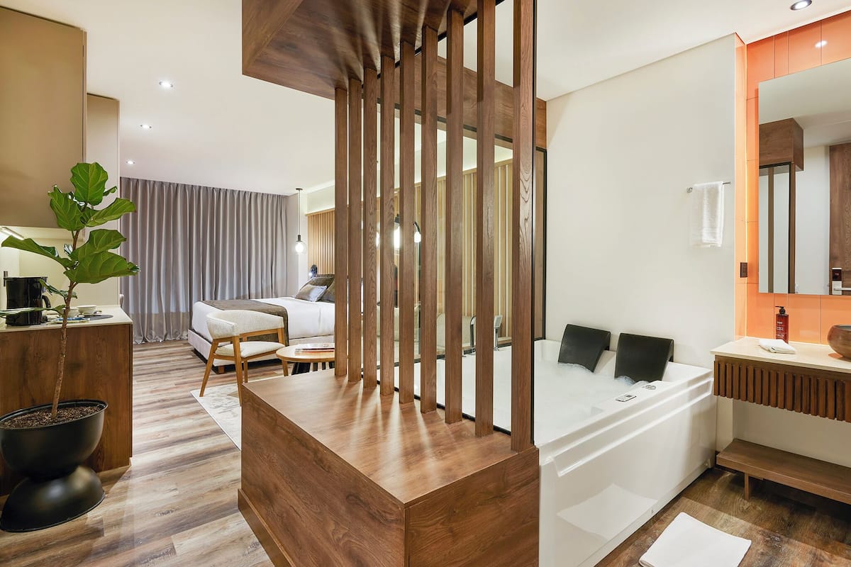 Una habitación de hotel moderna con un escritorio de madera, una planta grande en maceta, un sofá, una cama con cabecera blanca y un baño con bañera y espejo.