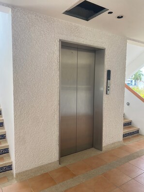 Een lift met een gestructureerde wand, een metalen hendel en een ventilatieopening aan het plafond.