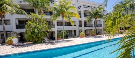 A poolside area with a clear blue pool, palm trees, and a white building with balconies.