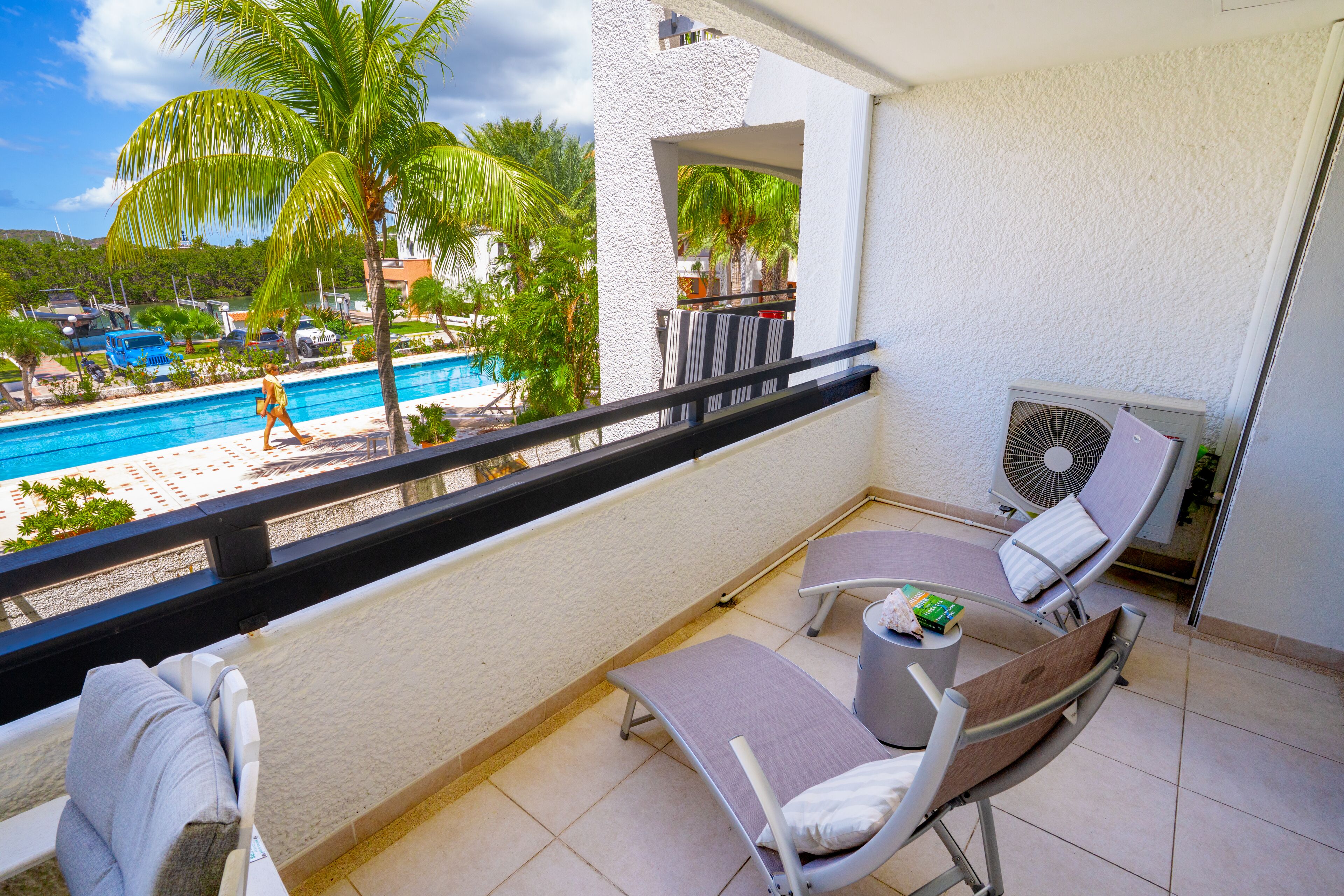 A balcony with two lounge chairs, a small table, and a fan. A pool and palm trees are visible in the background.