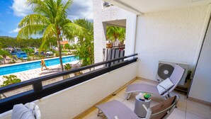 A balcony with two lounge chairs, a small table, and a fan. A pool and palm trees are visible in the background. - Simpsonbay Yachtclub apartment with pool (Cole Bay)