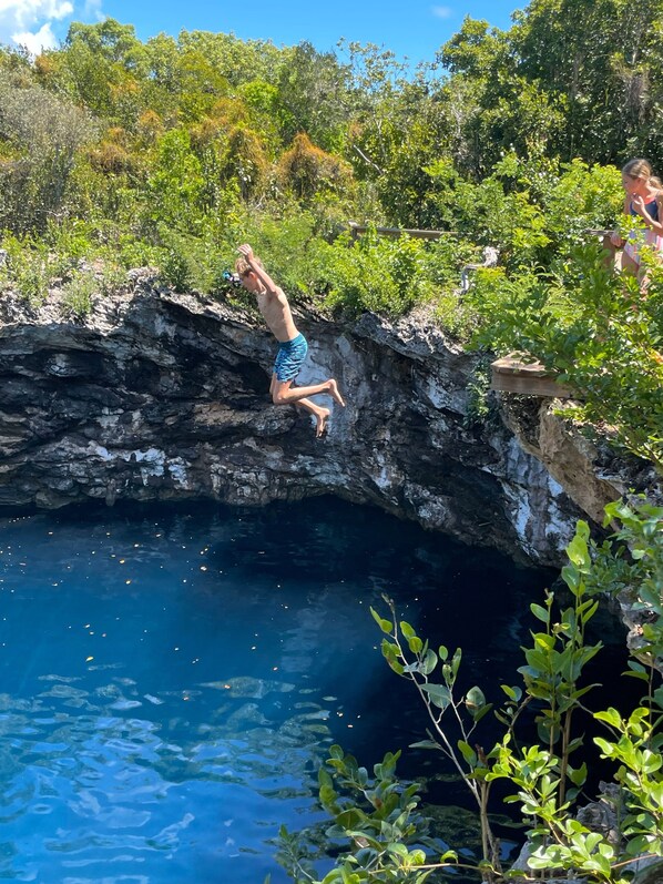 Una persona salta desde un acantilado rocoso a una piscina azul cristalina rodeada de vegetación.