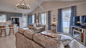 A spacious living room with a large sofa, a coffee table, and a television. There is a kitchen area with a refrigerator and a dining table with chairs. The room has large windows with curtains and a ceiling fan. - Gone Fishing on Lake Wateree (Liberty Hill)