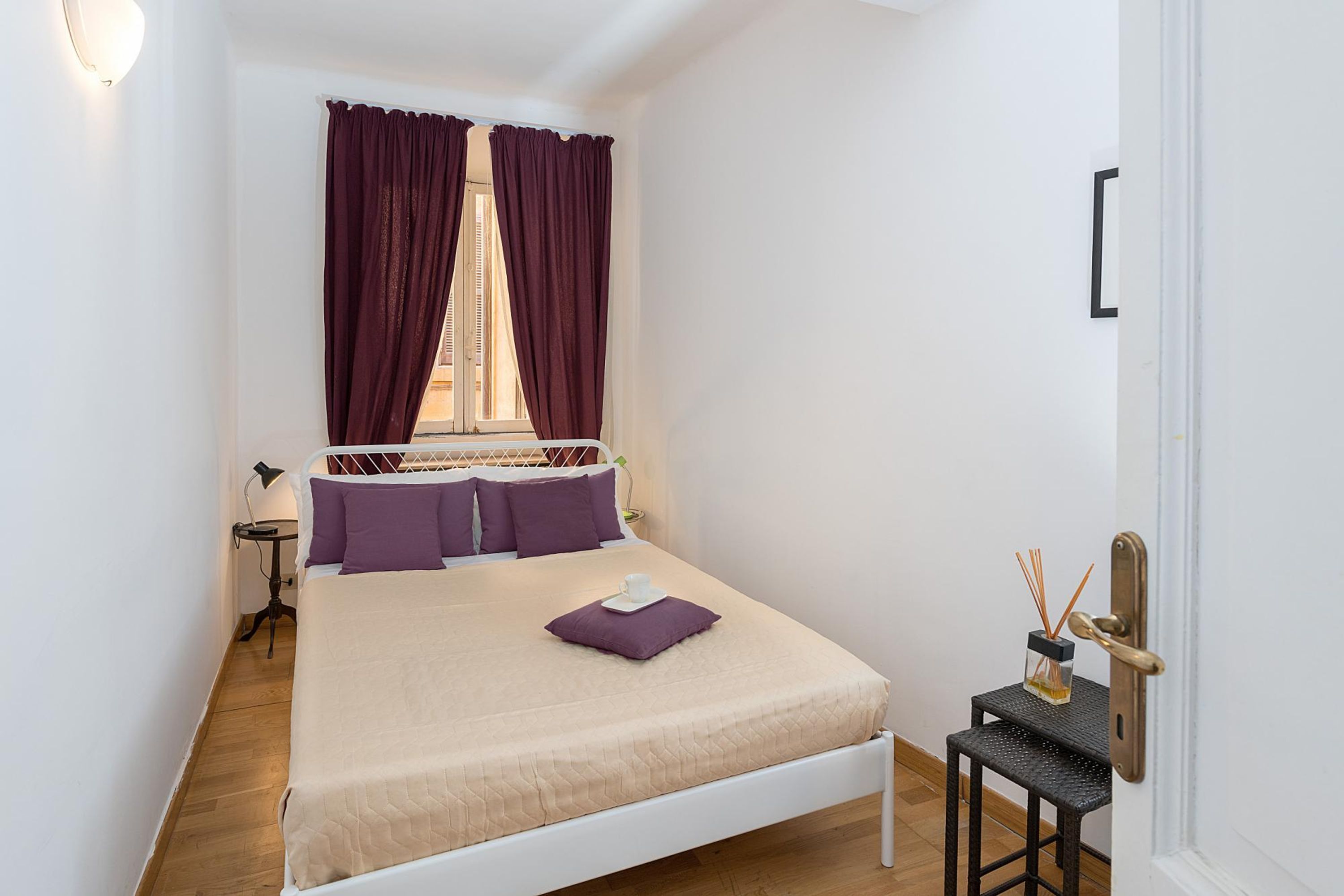 Un dormitorio ordenado con una cama, una mesita de noche, una pequeña mesa con un candelabro y una ventana con cortinas.