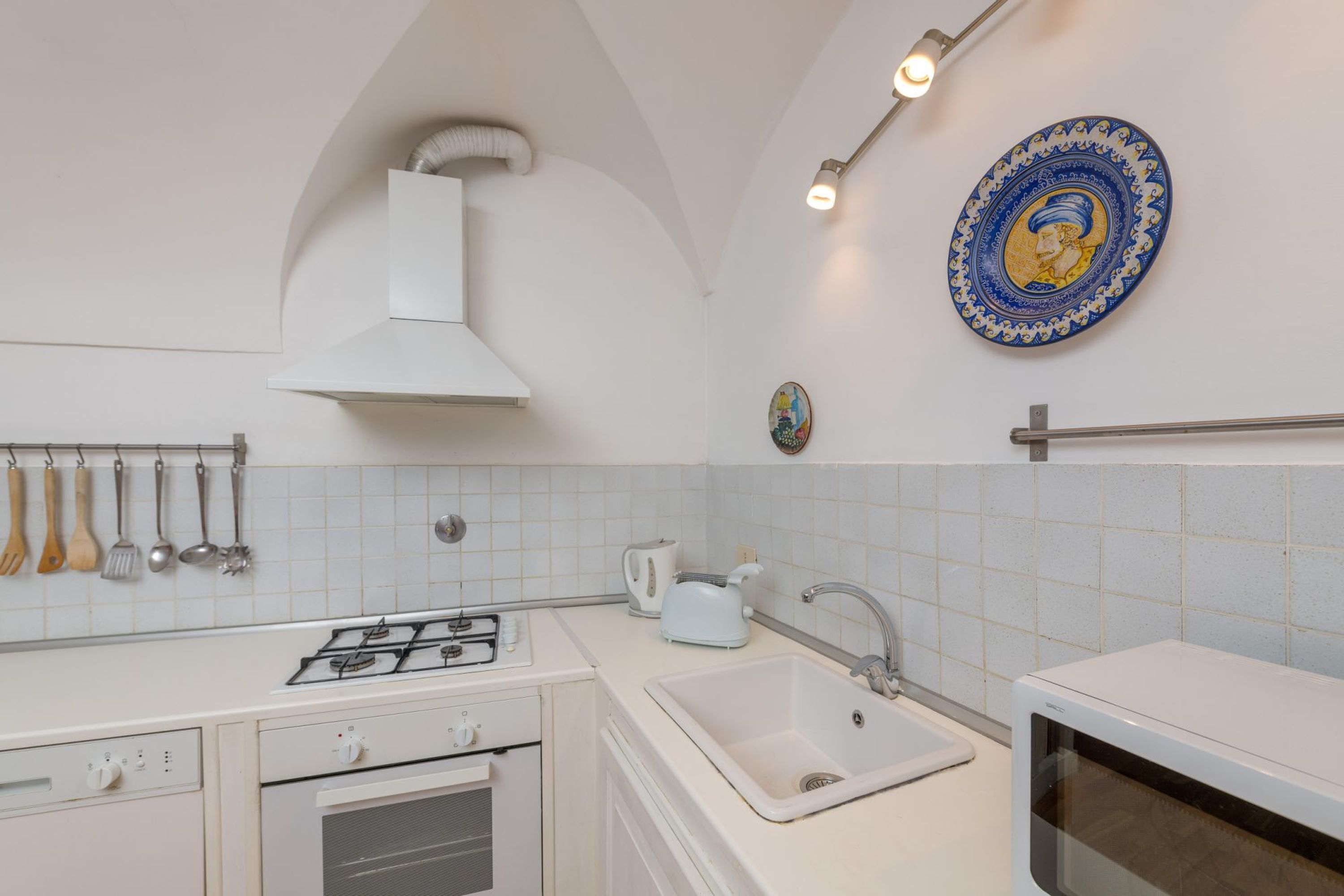 Une cuisine avec des meubles blancs, une cuisinière à gaz, un évier et un four à micro-ondes. On y voit des ustensiles de cuisine suspendus et une assiette décorative accrochée au mur.
