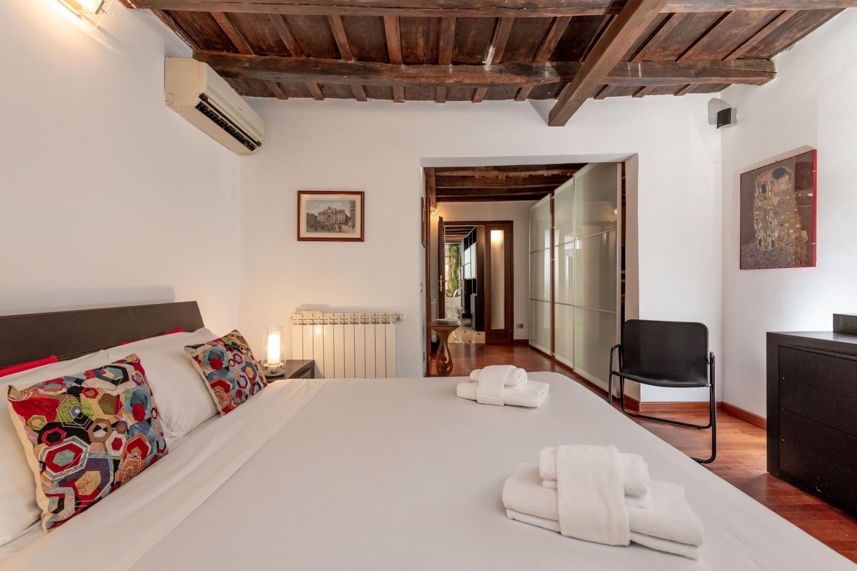A hotel room with a large bed, a wooden ceiling, a radiator, a chair, and a painting on the wall.