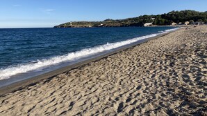 A sandy beach with gentle waves, a clear sky, and a distant hill with buildings.