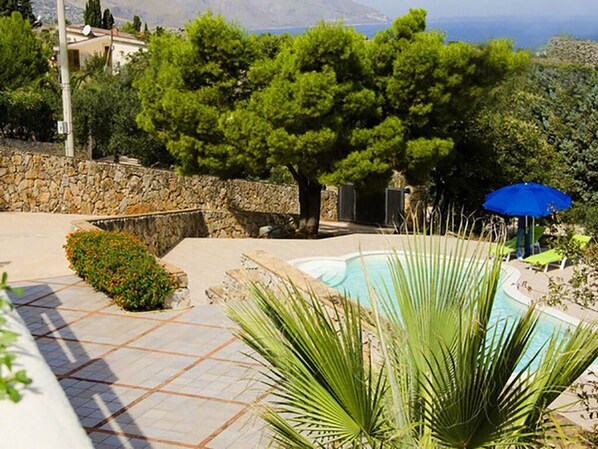 A pool area with a stone wall, a tree, and a blue umbrella. - Villa with swimming pool with spectacular sea views and Mediterranean vegetation (Sicilia)