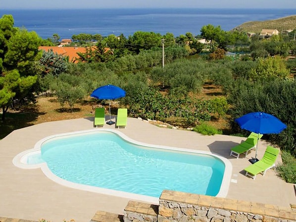 A swimming pool with two green lounge chairs and blue umbrellas, surrounded by greenery and a view of the sea.