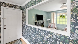 Ein Zimmer mit blumengemustertem Wandpapier, einer weißen Tür, einem Spiegel, der ein Fenster mit Vorhängen reflektiert, und einer Lampe.