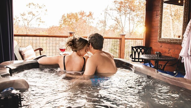 Een stel in een jacuzzi met een houten terras en uitzicht op bomen.