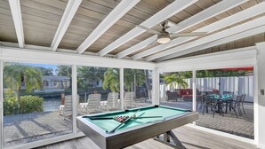 Una mesa de billar en una habitación luminosa con grandes ventanales, un ventilador de techo y mobiliario exterior visible a través del cristal.