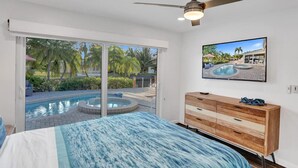 Un dormitorio con una cama, un aparador de madera, un ventilador de techo, un televisor en la pared y un ventanal corredizo que da a una zona de piscina.