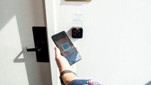 A hand holding a smartphone with a QR code, approaching a door with a room entry sign and a keycard reader.