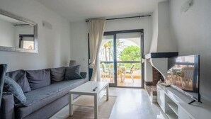 Een woonkamer met een bank, salontafel, televisie en een schuifpui naar een buitenruimte met stoelen.