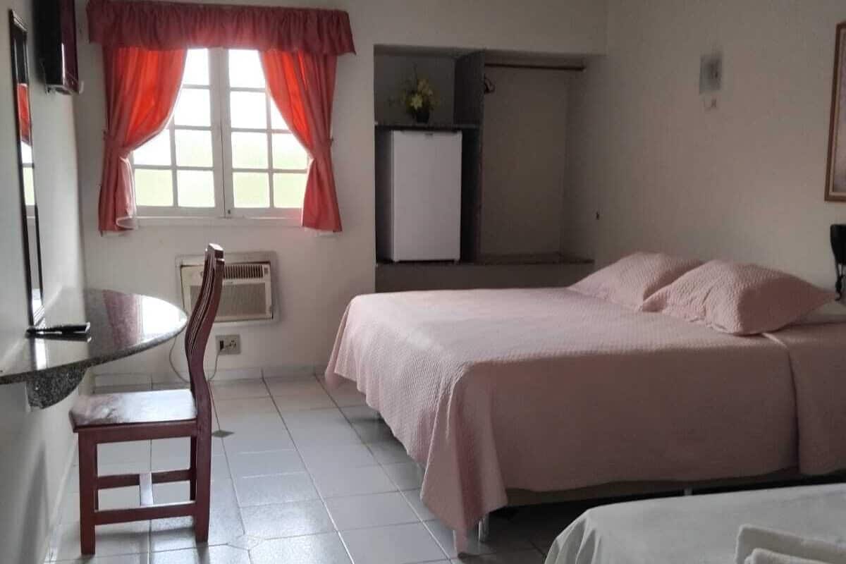 Een kleine, schone kamer met twee bedden, een bureau en een stoel. Er is een raam met gordijnen, een radiator en een klein kastje.