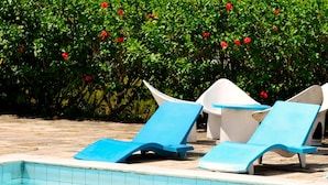 Un espace piscine avec des chaises longues bleues, une piscine carrelée et, au fond, un bâtiment dont le toit est également recouvert de tuiles.