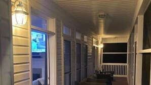 Un porche cubierto con un banco de madera y una silla, un televisor montado en la pared y una ventana con persianas.