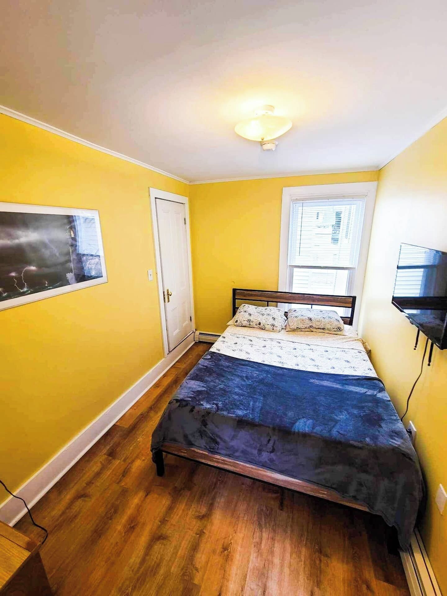 Un dormitorio con cama, puerta, ventana y televisor montado en la pared.