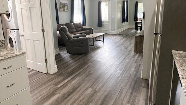 A modern living room with a grey sofa, a coffee table, and a dining area with a table and chairs. There is a kitchen area with a refrigerator and a washing machine. - Triple Play Apt 2 - All Star Baseball Rentals (Oneonta)