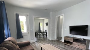 A modern living room with a sofa set, a coffee table, a TV, and a dining area with chairs and a table. - Triple Play Apt 2 - All Star Baseball Rentals (Oneonta)