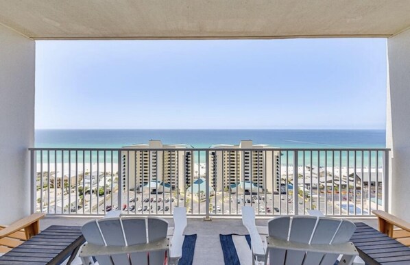 Een balkon met twee stoelen en een tafel, met uitzicht op een strand, gebouwen en een heldere lucht.