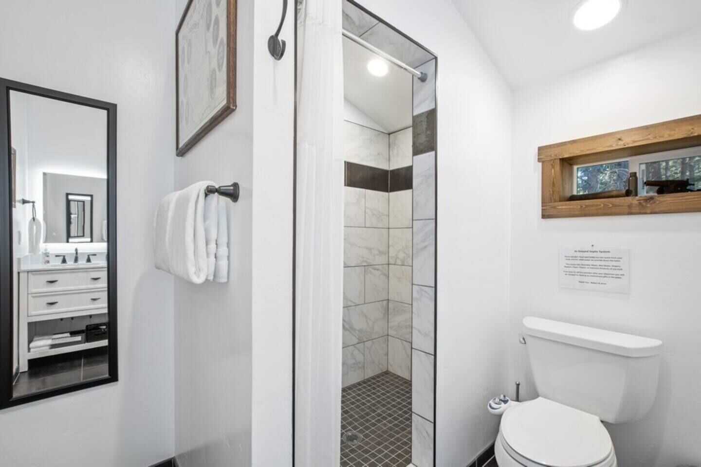 Un baño moderno con ducha envuelta en vidrio, inodoro blanco y estante de madera sobre el lavamanos.