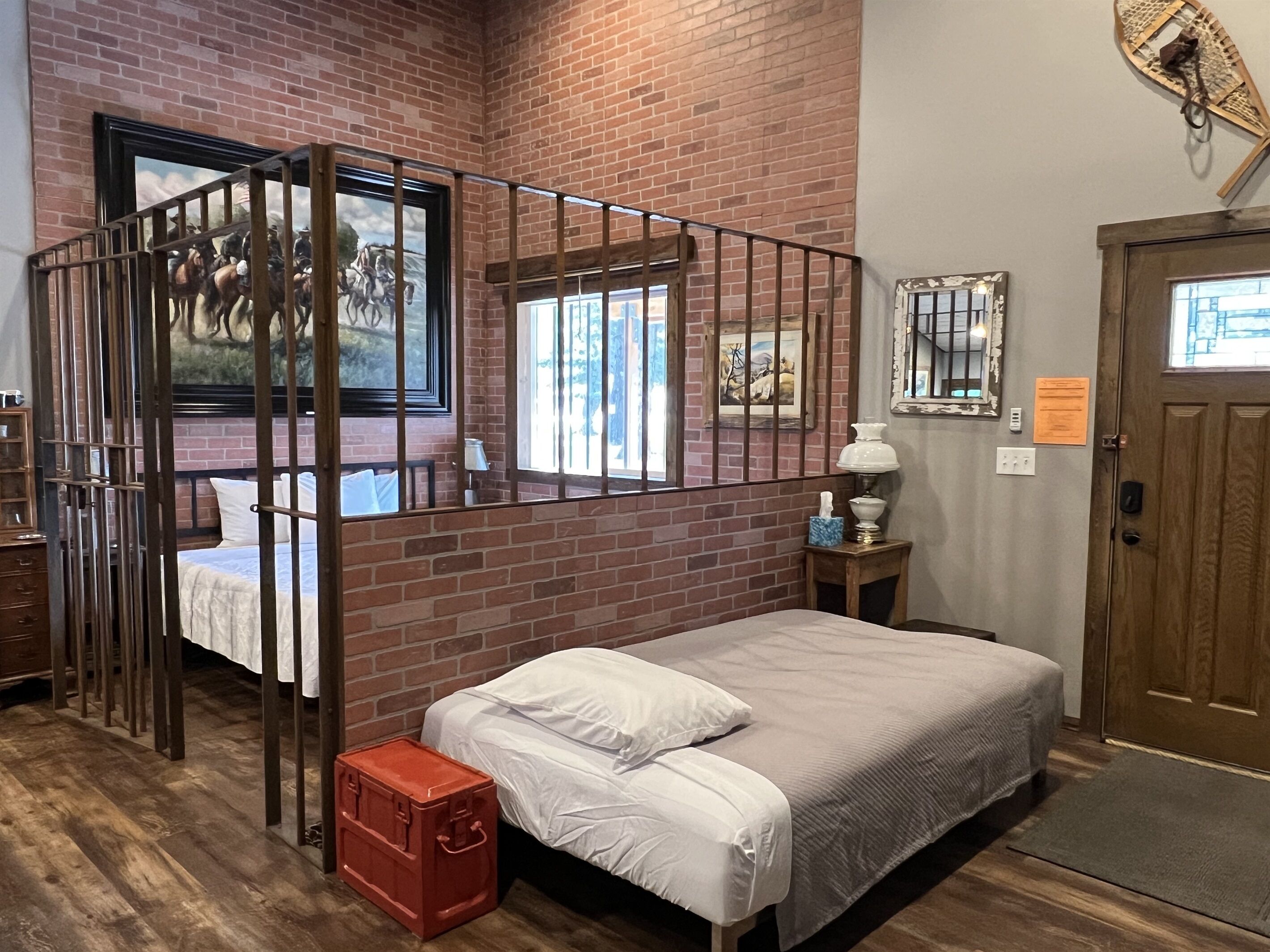 A room with a brick wall, a bed with white bedding, a wooden door, and a small table with a lamp.