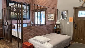 A room with a brick wall, a bed with white bedding, a wooden door, and a small table with a lamp.