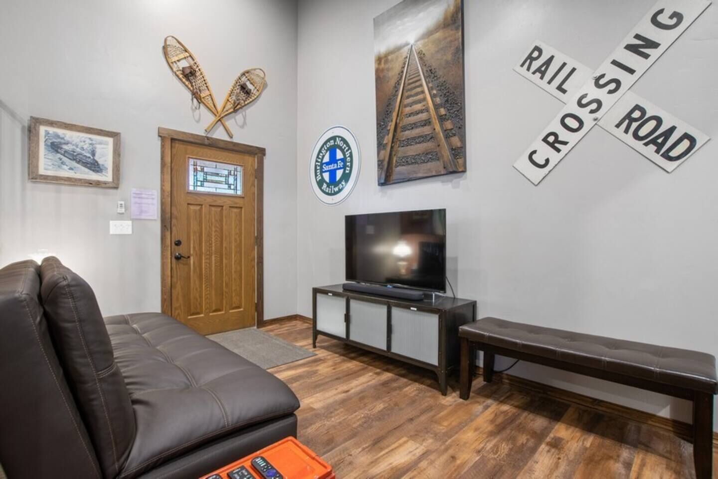 Un salon comprenant un canapé en cuir, un téléviseur posé sur un meuble, un banc en bois et des éléments de décoration murale, notamment un panneau indiquant un passage à niveau et le logo du chemin de fer Santa Fe.