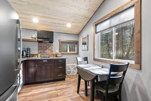 Una cocina con armarios de madera, un refrigerador, una mesa de comedor con sillas y un ventanal con vistas a árboles.