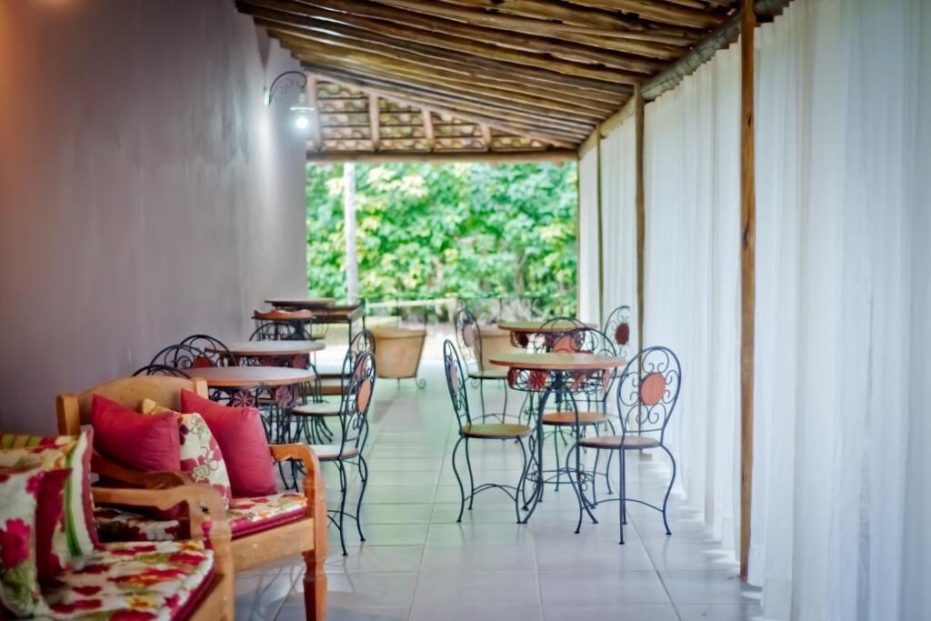 A covered outdoor seating area with tables and chairs, a wooden bench with cushions, and a view of greenery through the open sides.
