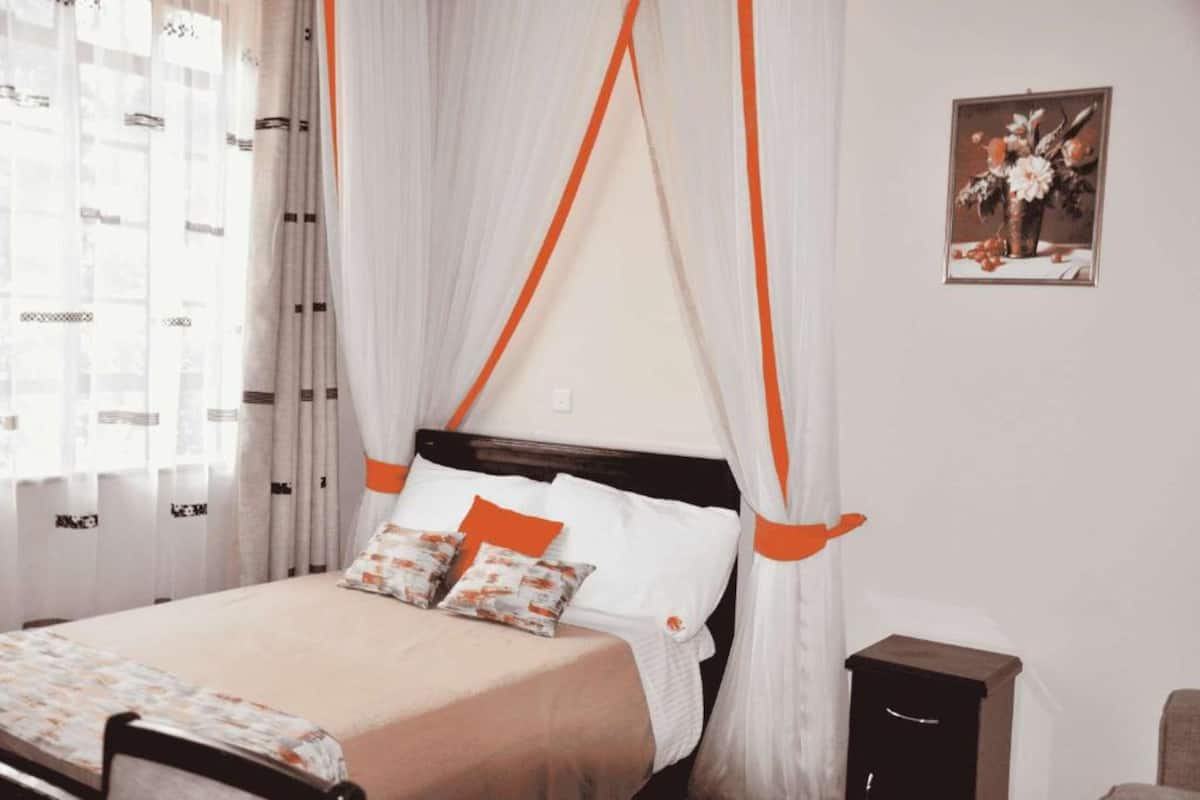 A bedroom with a bed, white curtains, a picture on the wall, and a bedside table.