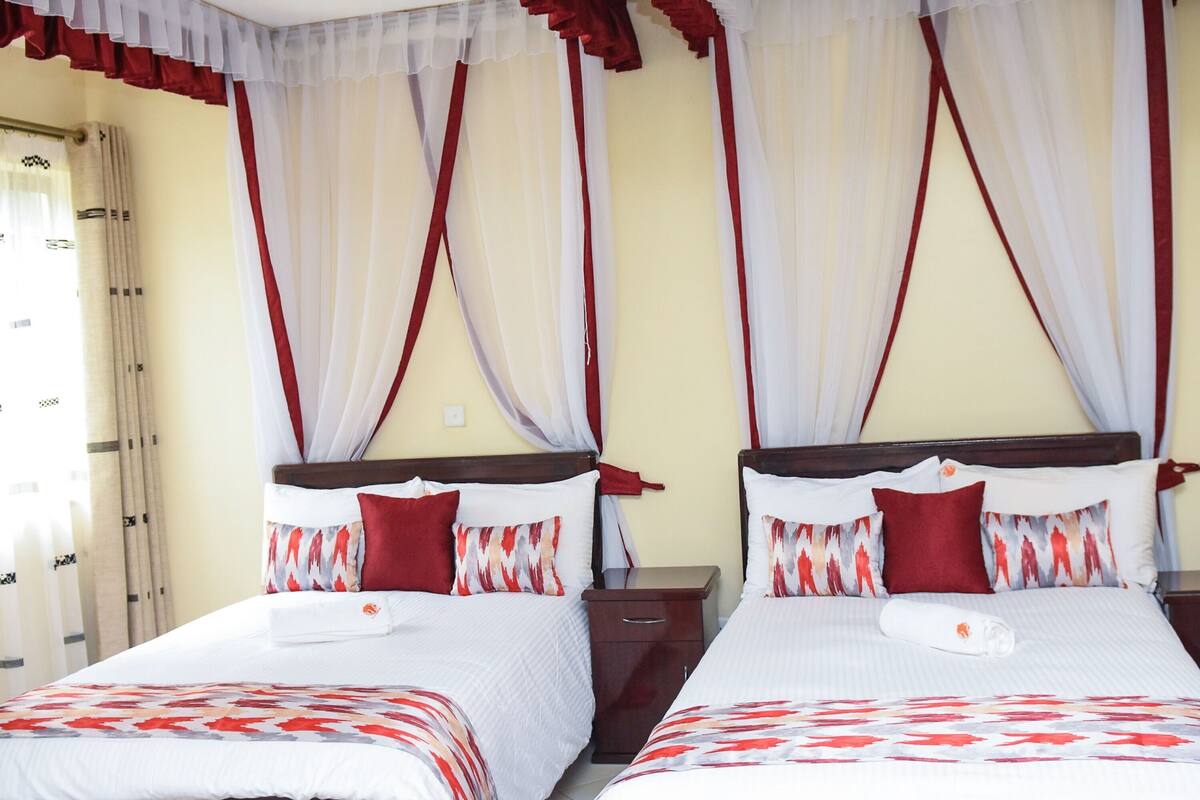 A hotel room with two beds, each with a white bedspread and red pillows, and sheer curtains with red trim.