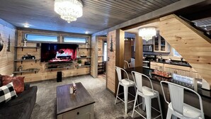Een gezellige woonkamer met een open haard, houten wanden en een televisie. Er staan witte stoelen en een kleine tafel.