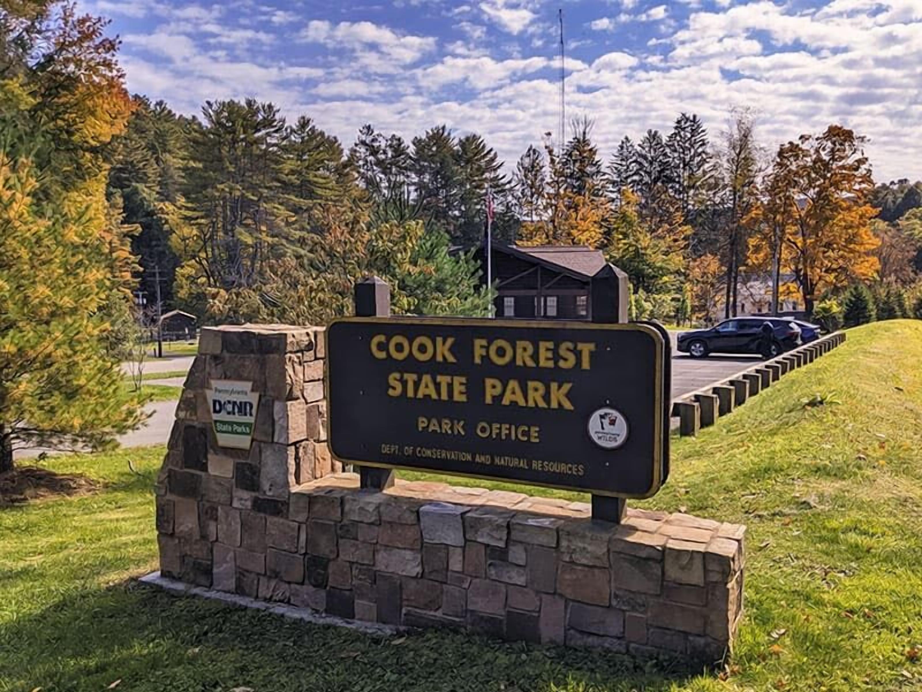 A sign for Cook Forest State Park with a stone base and a small building in the background.