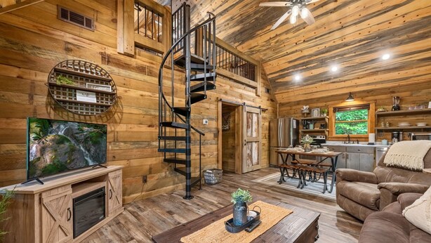 A rustic cabin interior with a wooden spiral staircase, a television on a wooden cabinet, a dining area, and a cozy seating area.
