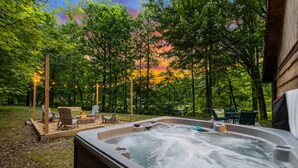 A hot tub surrounded by outdoor seating and string lights, set in a wooded area with a sunset in the background.