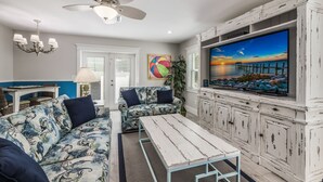A living room with a white ceiling fan, a TV on a white cabinet, a patterned sofa, and a table.