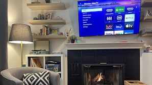 A living room with a fireplace, a television mounted above it, and a bookshelf with various items. - Family Friendly, Gated Yard for Pets, Outdoor Firepit! (Calgary)