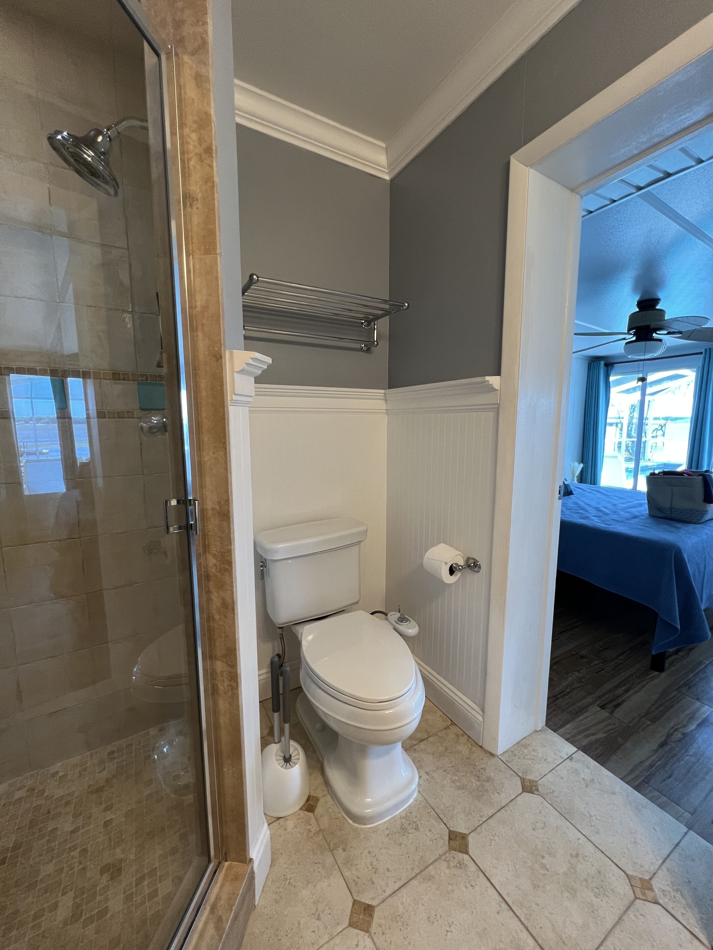 Een badkamer met een glazen douchecabine, een toilet en een blauw bed in de aangrenzende kamer.