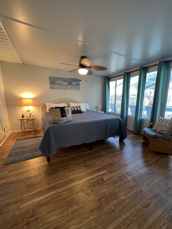 Un dormitorio con cama, ventilador de techo, mesita de noche, lámpara, ventana con cortinas y vistas al exterior.