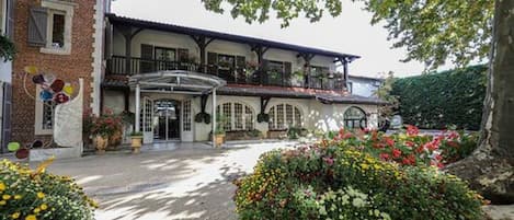 Edificio de dos plantas con balcón, vestíbulo cubierto y jardín lleno de flores.