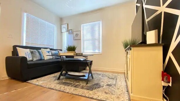 Un salon avec un canapé noir, une table basse et un tapis. Il y a deux fenêtres avec des stores, une étagère murale avec une plante et un élément décoratif géométrique noir et blanc sur le mur.