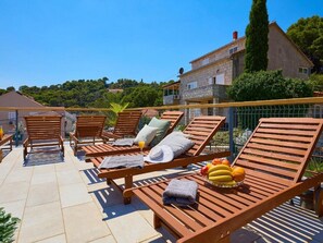 Una terraza con sillones de madera, una mesa con frutas y vistas a un edificio de piedra y árboles.