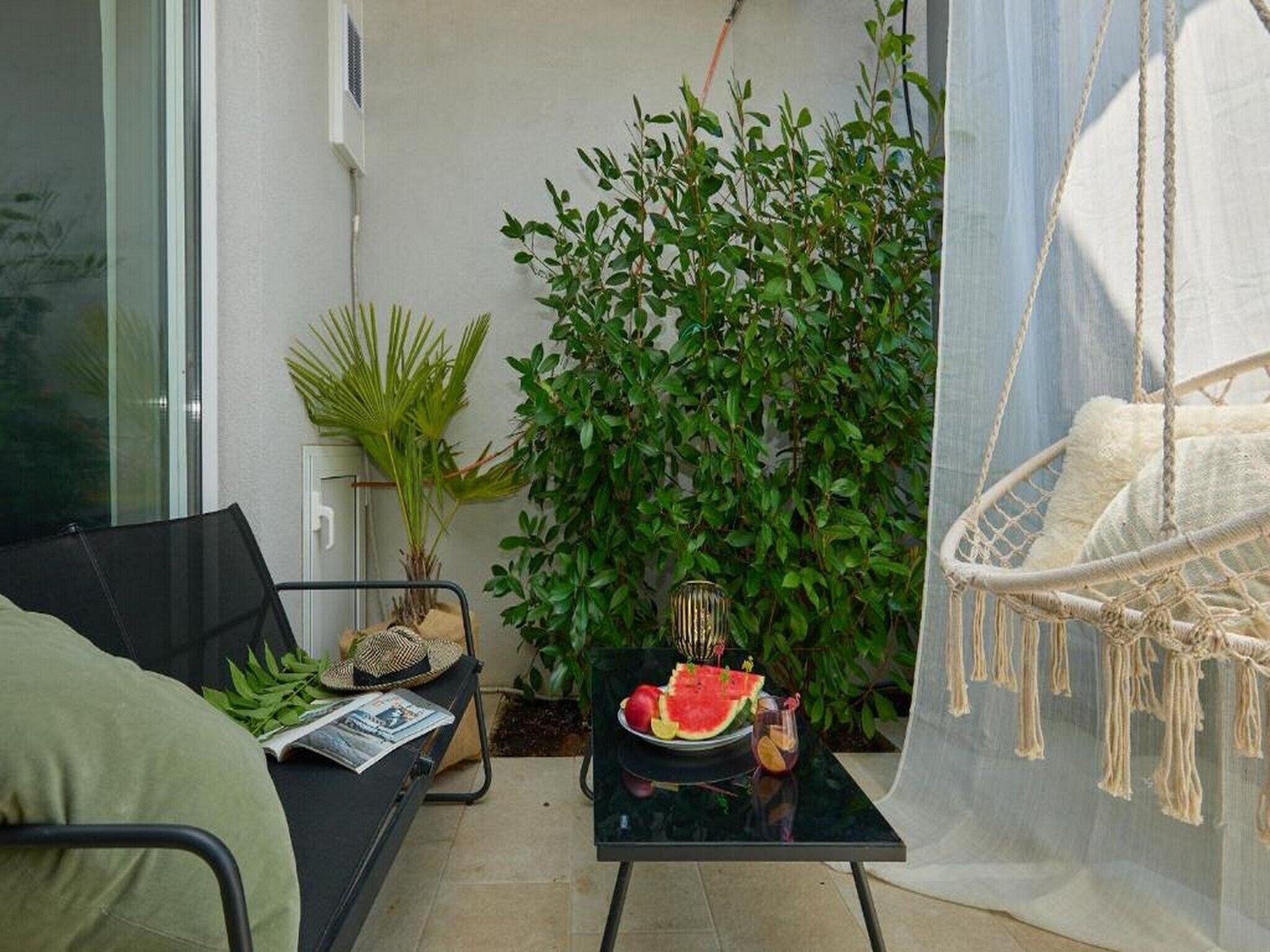 A small balcony with a black sofa, a glass table with a watermelon and drink, a hanging chair, and green plants.