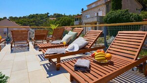 Eine Terrasse mit Holzliegen, einem Tisch mit Früchten und Blick auf ein Steinhaus und Bäume.