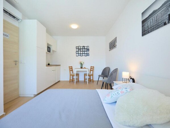 A compact hotel room with a bed, a small desk, a chair, a table, and a kitchenette.
