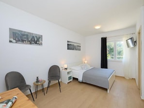 A modern bedroom with a bed, a desk, two chairs, a small table, and a TV mounted on the wall.